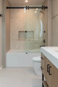 Modern bathroom with glass-enclosed bathtub, herringbone tile wall, wood vanity, and white countertop.