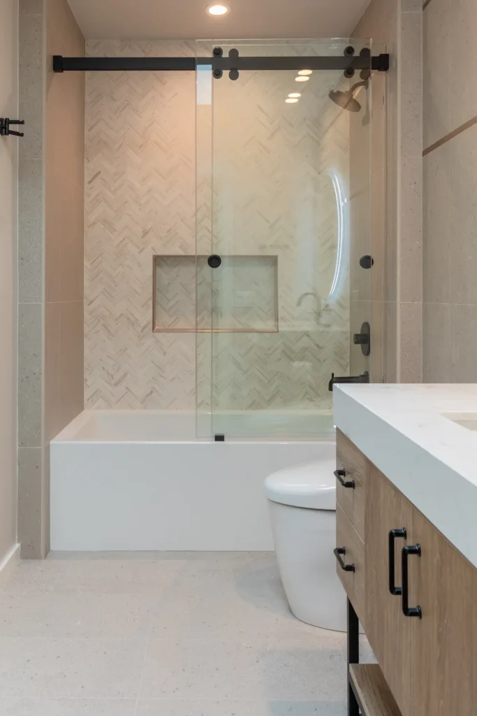 Modern bathroom with glass-enclosed bathtub, herringbone tile wall, wood vanity, and white countertop.