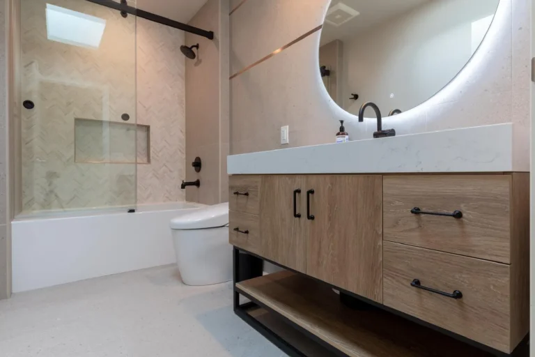 Modern bathroom with wood vanity, round mirror, black fixtures, and a glass-enclosed tub with tile surround.