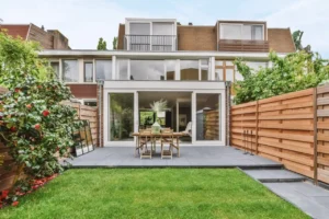Modern backyard with a wooden table, chairs, and landscaped greenery around the house.