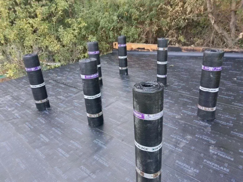 Flat roof surface covered in waterproof underlayment with rolled roofing materials standing upright on top.