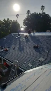 Roofers installing shingles on a roof with tools and materials visible, surrounded by trees.